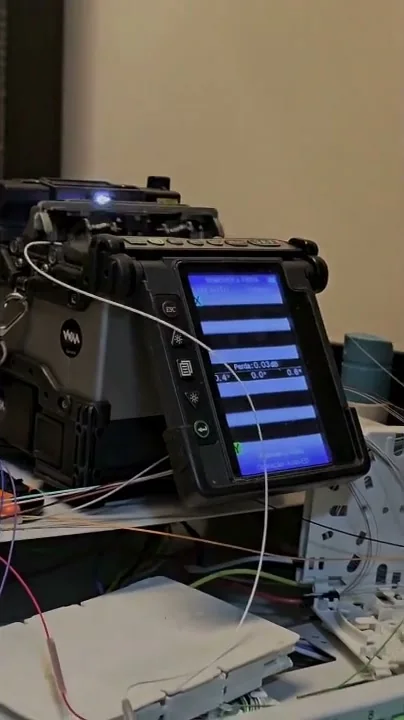 Máquina de fusão de fibra óptica sobre a bancada mostrando no visor a perda medida da emenda, com cordões ópticos e bandejas ao lado.
