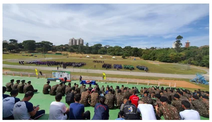 Público em uniforme militar e civis sentados na arquibancada acompanhando uma competição dos Jogos Militares em campo aberto na EsPCEx.